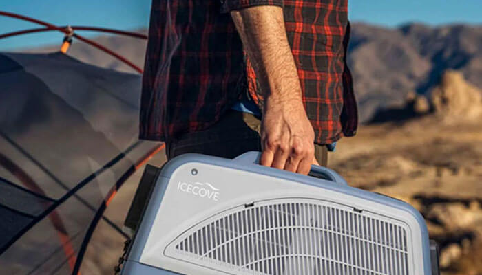 A portable air conditioner - tycoonstory | tycoonstory media Person in a red plaid shirt carrying a white portable cooler labeled icecove beside a tent at a rocky desert campsite.