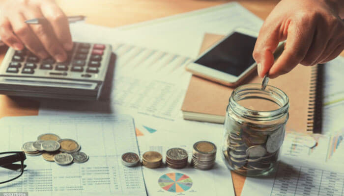 Budget and set expectations - tycoonstory | tycoonstory media Two hands counting coins into a glass jar on a desk cluttered with a calculator, charts, a notebook, and a smartphone.