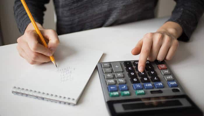Calculation basics with quick examples - tycoonstory | tycoonstory media Person writes on a notebook with a pencil while using a calculator on a white desk, showing a study or accounting scene.