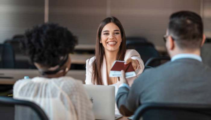 Create a pathway to citizenship 1 | tycoonstory media Smiling businesswoman at a desk hands a card to a client across a laptop in a modern office.
