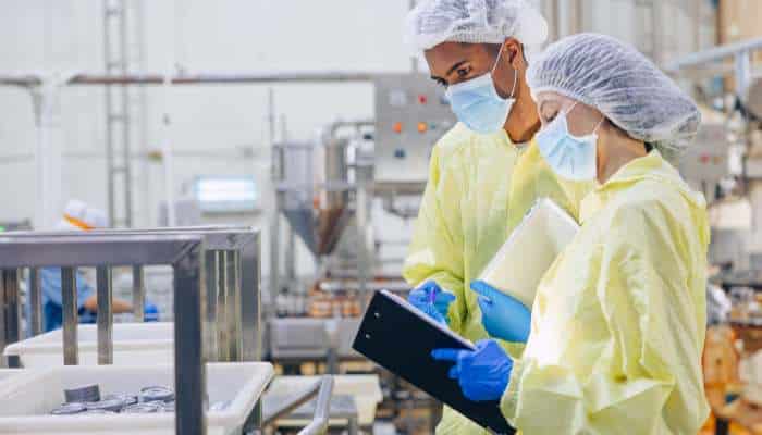 End-to-end processing solutions for modern food production 1 | tycoonstory media Two workers in yellow protective gowns, masks, and caps review a clipboard in a manufacturing facility.