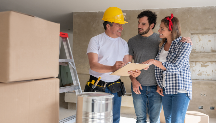 Home remodeling business | tycoonstory media Construction worker in a yellow hard hat discusses plans with a couple in a new-build room, holding a clipboard and a tablet.