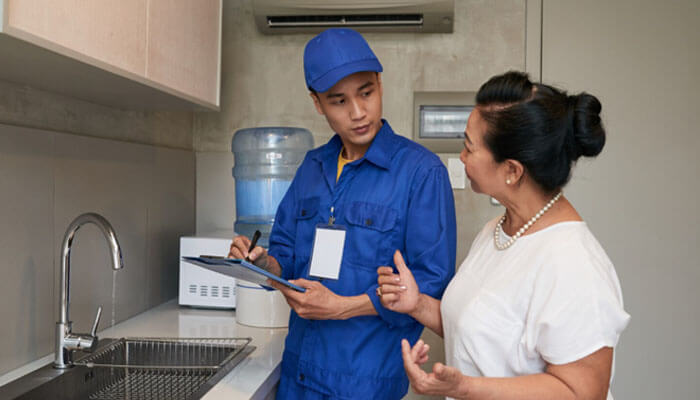 Check installation, maintenance, and lifespan - tycoonstory | tycoonstory media Delivery worker in blue uniform explains something on a clipboard to a woman in a white blouse in a kitchen area.