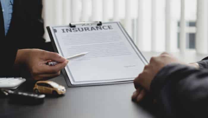 How a lawyer estimates your pi settlement amount 1 | tycoonstory media Two people sign an insurance contract on a clipboard with a pen, a small car model nearby suggesting auto insurance.