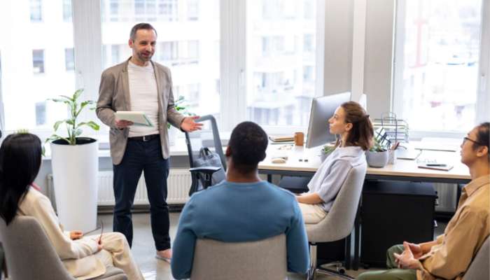 How a leadership coach helps you become a better decision maker 1 | tycoonstory media Manager presenting a project plan to a diverse team in a bright office meeting space, gesturing with a tablet in hand.