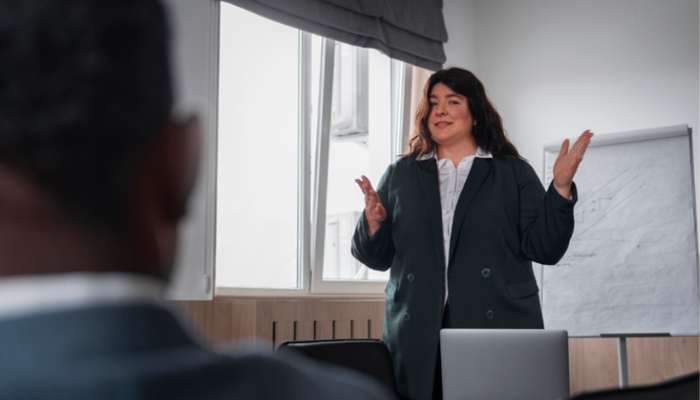 How a leadership coach helps you become a better decision maker 2 | tycoonstory media Female presenter in a blazer speaks to a room, gesturing with hands as she presents beside a flip chart near a window.