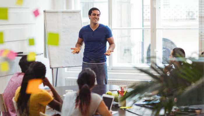 How a leadership coach helps you become a better decision maker 3 | tycoonstory media Man giving a presentation to a diverse team in a bright meeting room with post-it notes and a whiteboard nearby.