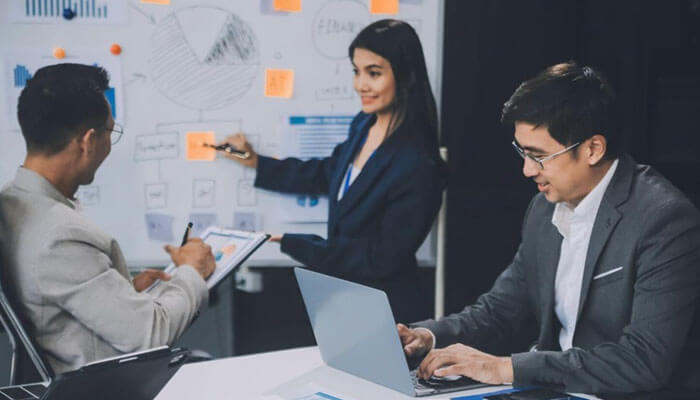 How headspin addresses modern testing challenges - tycoonstory | tycoonstory media Team in a modern meeting room; one woman presents charts on a whiteboard while two men discuss and take notes on a laptop and tablet.