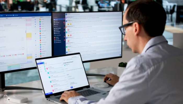 How to measure billable hours for agencies and freelancers 1 | tycoonstory media Man with glasses sits at a desk with three monitors and a laptop, monitoring chat and emails in a busy office. (informative)