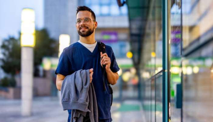 In which states travel nurses are finding the most work right now 1 | tycoonstory media Male nurse in blue scrubs walking outside a glass-front building, carrying a gray jacket and a backpack.