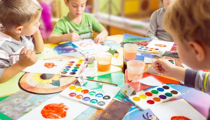 Innovative kindergarten lesson ideas to inspire young minds | tycoonstory media Children sit around a colorful table painting with watercolors and brushes, papers and cups of paint spread out in front of them, engaged in arts and crafts.