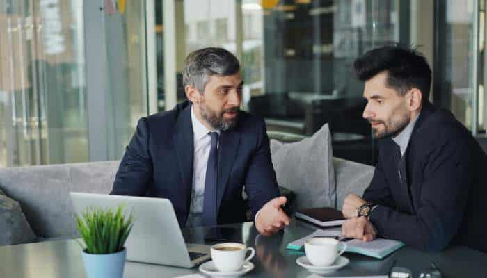 Integration is where budgets go to die | tycoonstory media Two men in suits discuss at a glass-walled office table, with a laptop, notebooks and coffee cups nearby — a professional meeting.