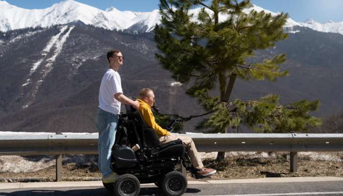 Manual vs electric wheelchairs how to choose the right one | tycoonstory media Man in a white shirt pushes a motorized wheelchair along a road with snow-capped mountains in the background.