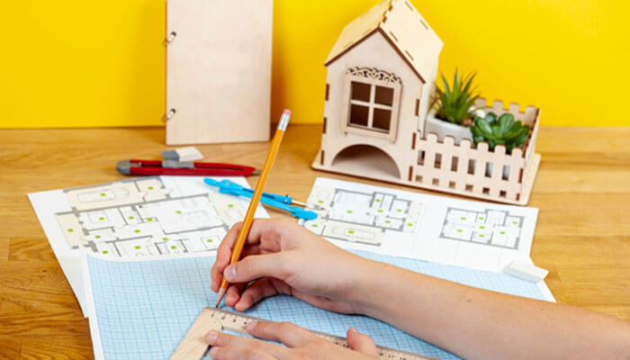 Map your home by zones and quick wins - tycoonstory | tycoonstory media Person draws on blueprints with a pencil and ruler on a wooden desk, a wooden house model and drafting tools nearby