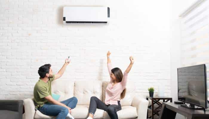 Mini split zoning | tycoonstory media Two people on a white sofa cheer with arms raised under a wall-mounted air conditioner in a bright, modern living room.