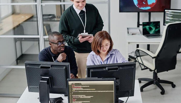 Network and failure simulation - tycoonstory | tycoonstory media Three colleagues collaborate at a desk with multiple monitors in a bright office; a woman types on a laptop while two men review screens and a supervisor stands with a tablet.
