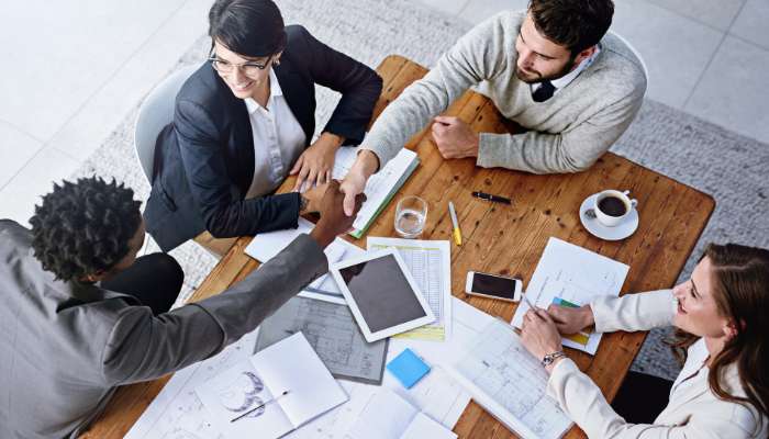 Offshoring is how smart us businesses are staying ahead 1 | tycoonstory media Overhead view of a diverse four-person team at a wooden table, shaking hands amid documents, a tablet, and coffee cups, signaling agreement.