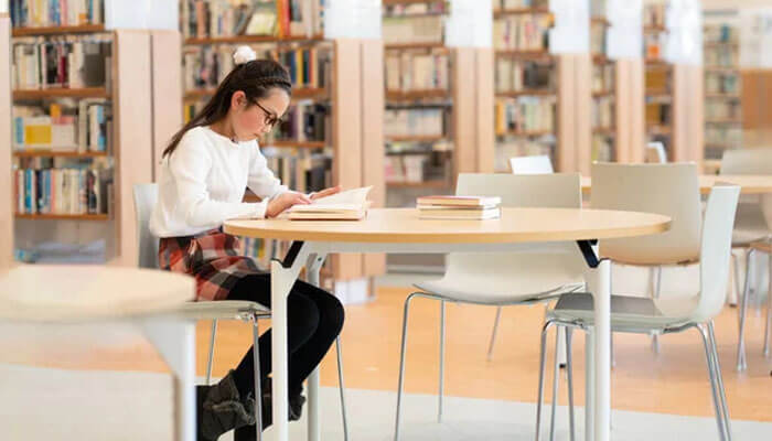 Reading the room in mcmullen county - tycoonstory | tycoonstory media Young woman reading a book at a round table in a library, with tall bookshelves in the background.