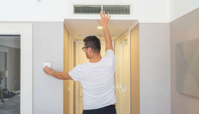 Signs your home is not performing as efficiently as it should 2 | tycoonstory media Man in a white t-shirt and glasses reaches up to touch a ceiling vent above a double glass elevator door in a bright hallway.