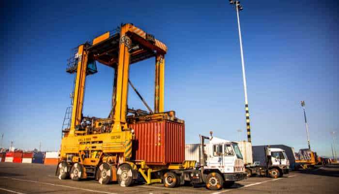 Straddle carrier price 1 | tycoonstory media Straddle carrier price analysis with a container handling machine operating at a busy shipping terminal.