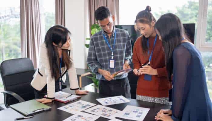 The hidden factors behind local search rankings 1 | tycoonstory media Four colleagues standing around a conference table, examining printed charts and documents in a bright office setting with badges on lanyards.