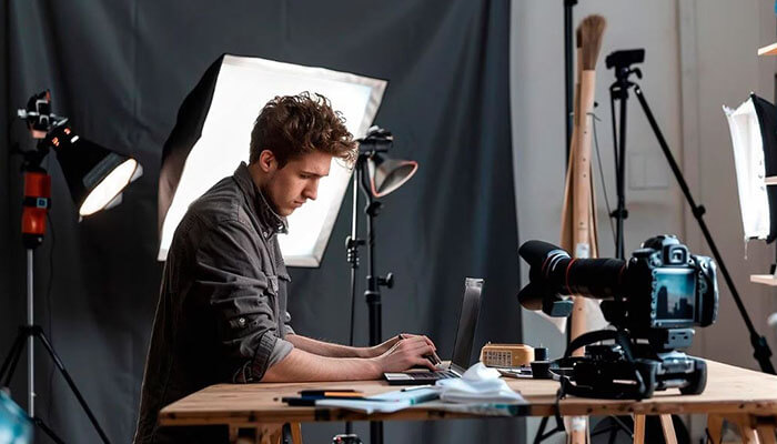 The marketing problem that comes after the creative one - tycoonstory | tycoonstory media Man sits at a wooden desk editing on a laptop in a photography/video studio with professional lighting and equipment nearby.