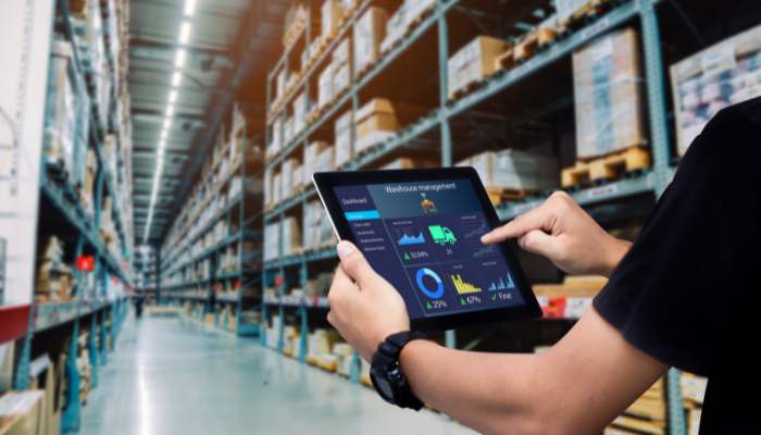 Top 5 benefits of upgrading your warehouse management software 1 | tycoonstory media Person in a warehouse uses a tablet showing analytics charts on a blue dashboard screen in a long storage aisle.