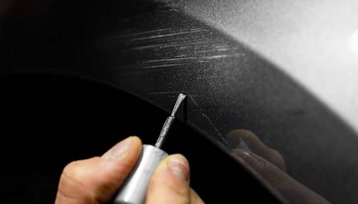 Touch-up paint for drivers 1 | tycoonstory media Close-up of a hand using a small rotary tool to scratch a dark curved surface on a vehicle panel.