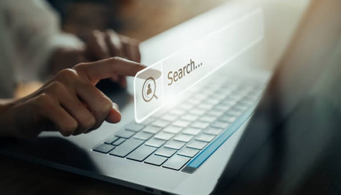 Untitled-1 | tycoonstory media Person typing on a laptop with a translucent floating search bar reading 'search... ' above the keyboard to suggest online searching