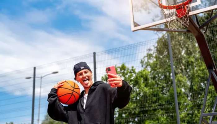 Video views are especially important for sports content | tycoonstory media Man in a black hoodie and cap takes a selfie while holding a basketball on an outdoor basketball court, hoop visible overhead.