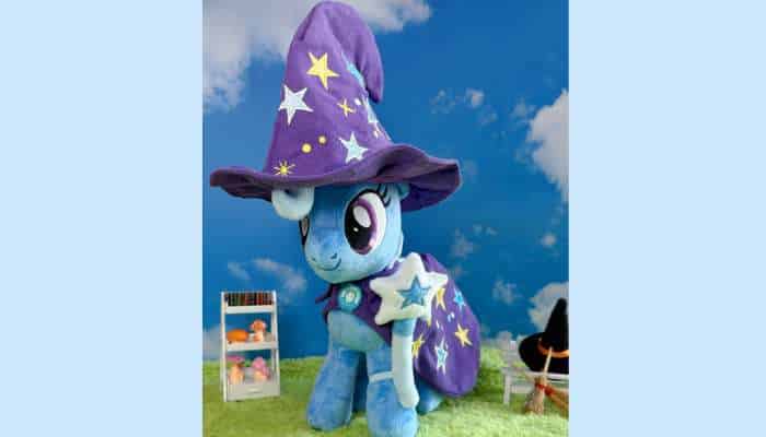 What makes symbiote studios trixie lulamoon deluxe plush stand out from standard plush toys 1 | tycoonstory media Rixie lulamoon deluxe plush wizard pony toy wearing purple star hat and cape in magical outdoor sky scene display