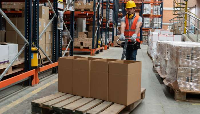 What to look for in a reliable logistics partner for your brand 1 | tycoonstory media Warehouse worker in a yellow hard hat and orange vest uses a pallet jack to move a pallet with four brown boxes in a busy warehouse.