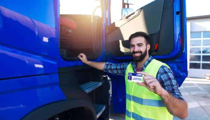 What your cdl class says about what you can drive | tycoonstory media Man in a high-visibility vest shows an id badge beside a blue semi-truck cabin with the door open, outdoors.