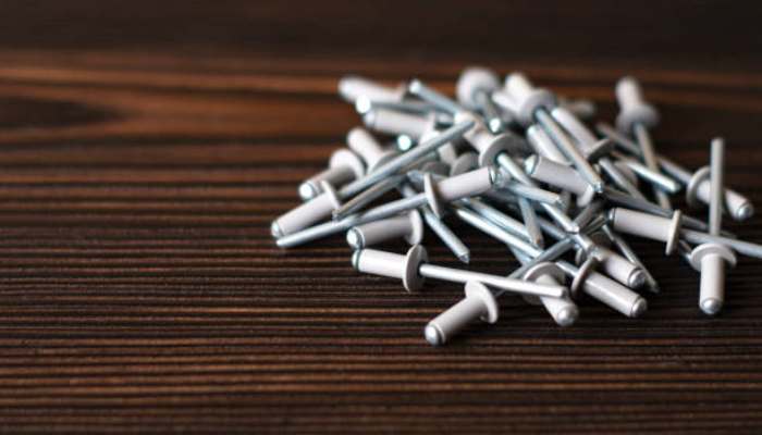 When to use a countersunk rivet for a flush surface connection 1 | tycoonstory media Pile of metal finish nails with white heads scattered on a dark wooden surface, ready for carpentry tasks.