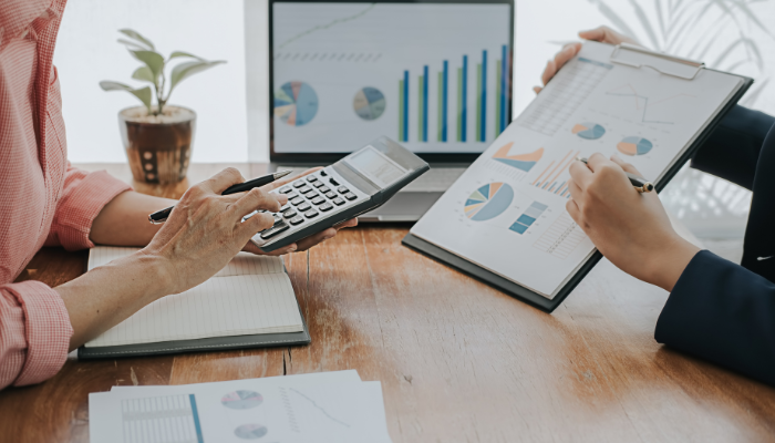 Why detroit is popular amongst top businesses 1 | tycoonstory media Two professionals review data: one uses a calculator while the other points to a clipboard filled with charts; a laptop on the table displays graphs.