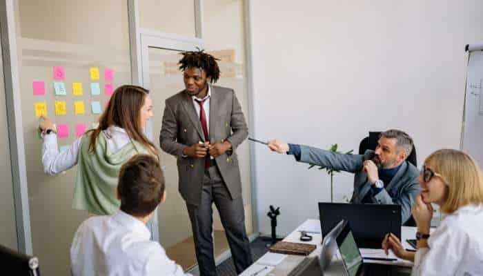 Why growing companies need analytics services | tycoonstory media Team brainstorming in an office: a woman places sticky notes on a glass wall as a suited man presents, others observe and discuss around a conference table.