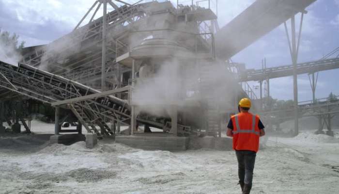 Why is silica dust monitoring important in the workplace 1 | tycoonstory media Worker in an orange safety vest and yellow hard hat walking toward a large industrial processing plant with conveyors and steam clouds around the equipment (construction/industrial site).