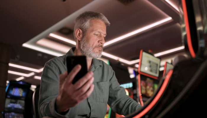 Why malaysian players are choosing vip2cuci casino malaysia over traditional platforms 1 | tycoonstory media Older man with gray beard looking at his phone while seated at a casino or arcade gaming machine area.