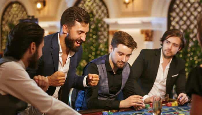 1cinta online casino 1 | tycoonstory media Four men in smart-casual attire sit around a casino table, smiling and placing bets as chips and cards are visible.