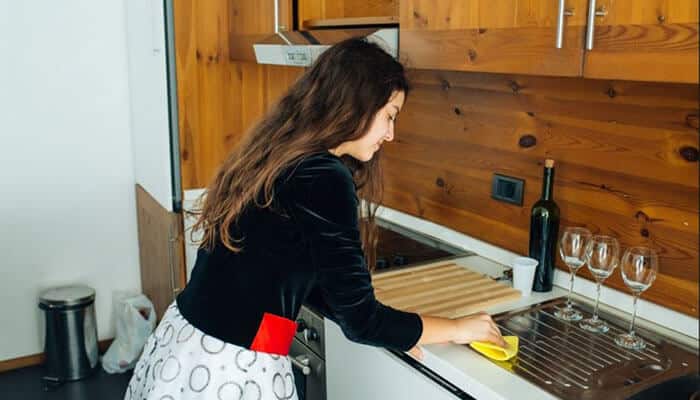 Reducing the frequency and costs of applying artificial sealants - tycoonstory | tycoonstory media Woman washing dishes at a stainless steel sink in a wood-toned kitchen, using a yellow sponge.