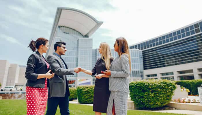 New off-plan luxury projects in dubai - tycoonstory | tycoonstory media Four businesspeople in professional attire stand outside a modern office complex; two women and two men are shaking hands to seal an agreement.