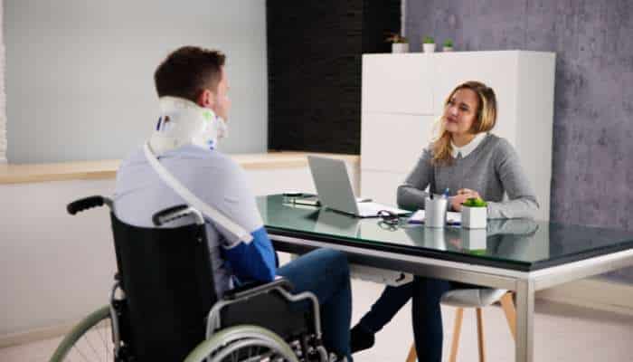 Answer engines and the future of how clients find law firms 3 | tycoonstory media Man in a wheelchair with a neck brace talks with a woman at a glass desk in a bright office, suggesting a consultation or interview.