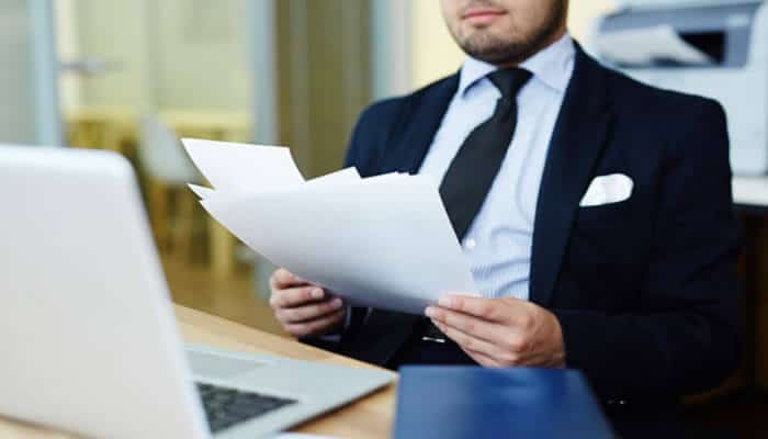 Submit required documents - tycoonstory | tycoonstory media Businessman in a suit reviewing documents at a desk with a laptop nearby.
