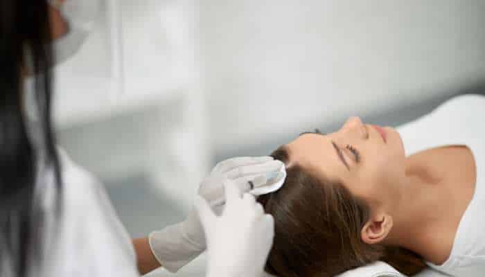 Choosing the right hair restoration clinic in melbourne 1 | tycoonstory media Woman lying on a treatment table with eyes closed as a clinician in gloves applies a cloth to her scalp.