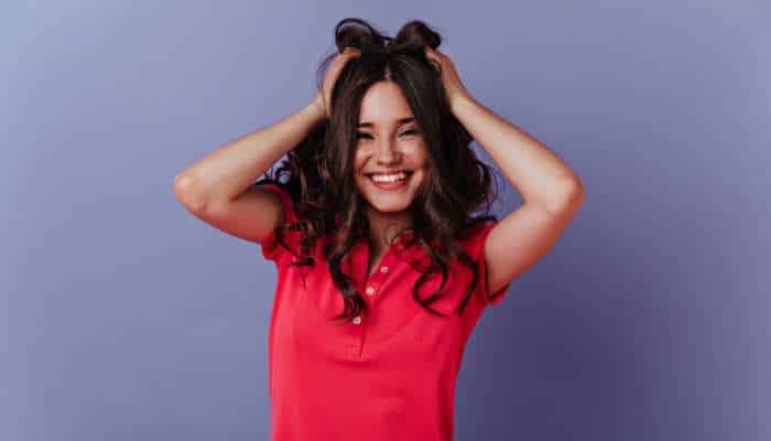 Choosing the right hair restoration clinic in melbourne 2 | tycoonstory media Young woman with long dark hair, wearing a red polo shirt, smiling with hands in her hair against a purple backdrop.