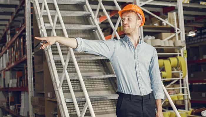 Why the routine rarely changes, and why that's a strength - tycoonstory | tycoonstory media Male worker in a blue shirt and orange hard hat points toward the left beside a metal staircase in a warehouse moment? Observing.