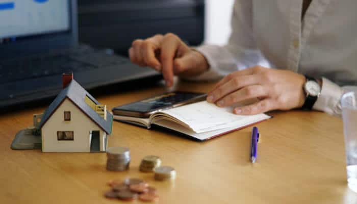 Borrower profile affecting home loan interest rate - tycoonstory | tycoonstory media Person in a white shirt uses a calculator and writes in a notebook on a desk; a miniature house and coins sit nearby.