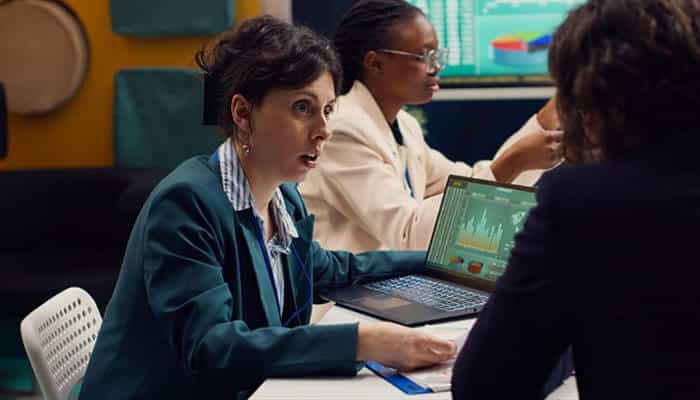 How cyber risk evaluation affects valuation and deal terms - tycoonstory | tycoonstory media Two professionals in a meeting discuss data, with a laptop showing chart graphs on the table in a modern office setting.