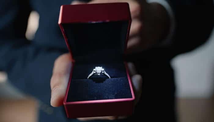 Gvftbhfgbn | tycoonstory media Diamond engagement ring in a red jewelry box with a dark blue interior, being held toward the camera by a person