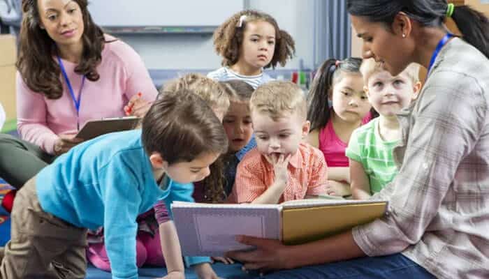 Supporting the hiring and training of trusted educators - tycoonstory | tycoonstory media Teacher reading to a diverse group of preschool children in a classroom implementing early childhood education frameworks, encouraging literacy, language development, emotional wellbeing, and inclusive early childhood education.